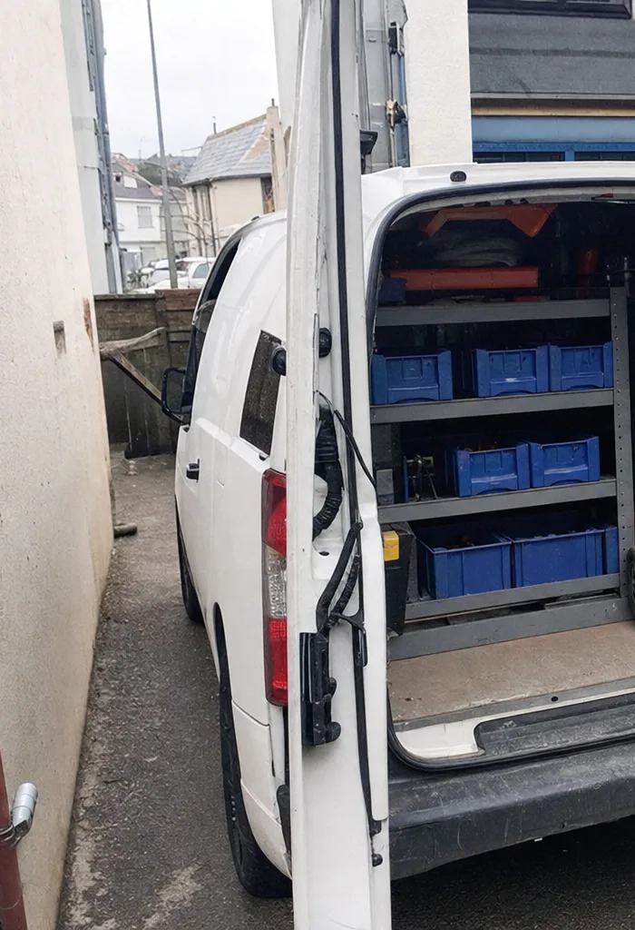 A white service van pulled up tight against a rendered wall in a narrow side passage, its rear door swung open on shelves of blue parts bins.
